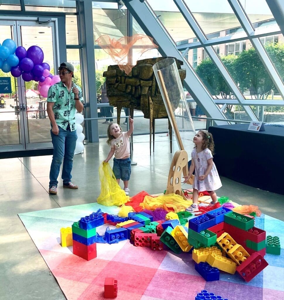 Two babies look up in the air at some balloons, a person is standing behind them watching them play.